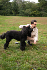 Young man on the park lawn playing with a royal black poodle
