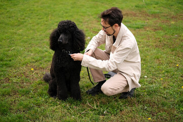 Young man on the park lawn playing with a royal black poodle