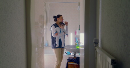 Mother brushing her teeth while holding her baby in the bathroom. baby looks around curiously,...