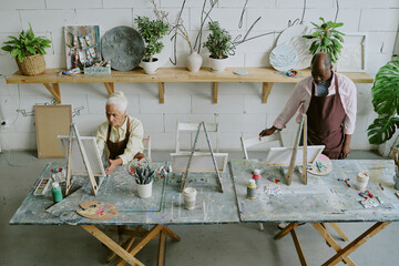 High angle shot of senior biracial artists working at work desk with paintings tools in retro light...