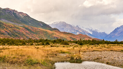This stunning landscape, located near El Chaltén in Patagonia, Argentina, features autumn colors, mountainous terrain, and a serene stream winding through the grassy fields below the Andes.
