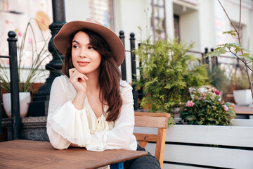 Portrait of beautiful young woman .in hat. Model at coffee shop. Female outdoors in sunny day. In white blouse. Cheerful and happy. Waiting for an order at terrace cafe, looks great