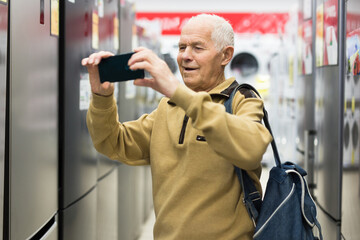 Elderly man taking photo of refrigerator in showroom of electrical appliance store