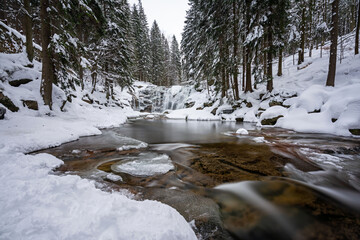 Fototapeta premium Captivating Winter Landscape: Frozen Waterfall Cascading into a Flowing River Surrounded by Snow-Covered Coniferous Trees - A Breathtaking Display of Nature's Icy Beauty