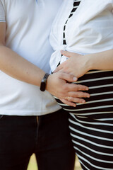 A pregnant woman in the arms of her husband. Summer sunny weather. A couple in love are hugging. Future parents. Young mom. Happy addition to the family. The concept of clothes for pregnant women.