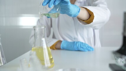 A scientist, wearing a lab coat and blue protective gloves, is pouring a yellow liquid from one beaker to another in laboratory, close up. Medicine and science concepts