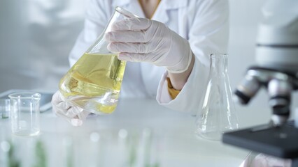 Microscope is near a scientist, wearing a lab coat and white protective gloves, and holding beaker with a yellow oily liquid in laboratory setting, close up. Medicine and science concept