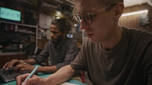 Medium shot of nerdy young Caucasian technician writing down specifications for new product while developing draft, and Asian male colleague typing on laptop computer
