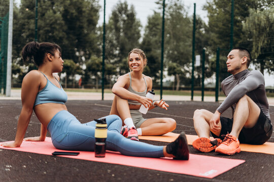 Diverse group of smiling attractive women and man sitting on yoga mat relaxing on the street