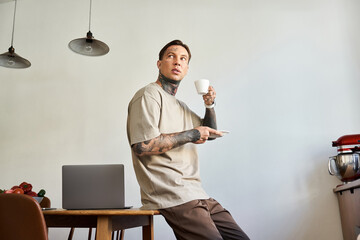 The young man sips coffee, contemplating while seated in a stylish kitchen workspace.
