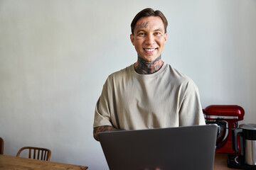 A cheerful young man with tattoos stands in a cozy home environment, engaging with his laptop.
