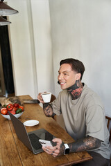 This young man joyfully sips coffee while engaged with his laptop in a warm, inviting space.