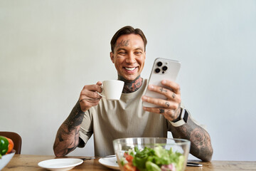 A cheerful young man is sipping coffee and smiling at his smartphone during breakfast at home.