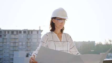 Woman architect, wearing safety helmets and glasses, is holding blueprints and inspecting a building site during sunrise or sunset, front view portrait. Architecture and engineering