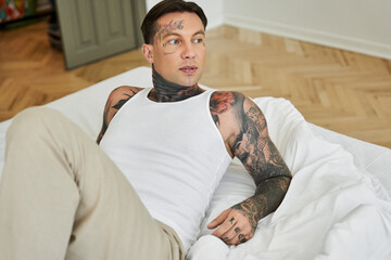 A handsome young man lounges on a bed, displaying intricate tattoos and a casual style.