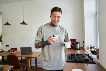 A young man stands in his kitchen, sipping coffee and smiling at his phone while relaxing.