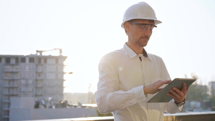 Male construction engineer or architect wearing white shirt and hard hat is using digital tablet while inspecting a building site at sunset, front view