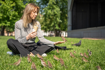 Contented woman communication with street sparrows while rest on urban lawn with ice cream. Smiling pleased female take break outdoor on grass in park eats sweetness watching sparrows begging for food