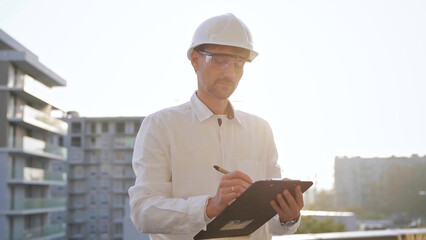 Man construction engineer wearing white shirt and hard hat is making notes on a clipboard while inspecting a building site at sunset, front view. Architecture and engineering concepts