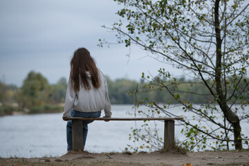A girl or a young woman sits with her back to us on a bench in front of the water of a lake or...