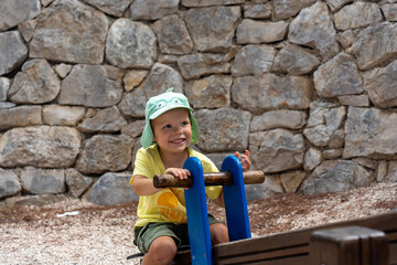 Happy child enjoying a playful ride on a seesaw in the park, wearing a cheerful green hat and a bright yellow t-shirt. Concept of active lifestyle, outdoor fun, and childhood adventures in nature.