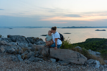 Mother and young son enjoying sunset on rocky hilltop with scenic sea and island view. Concept of family bonding, outdoor adventure, and nature appreciation.