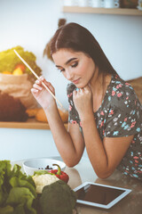 An attractive young dark-haired woman tastes a new recipe for a delicious salad mix while sitting at the table in sunny kitchen