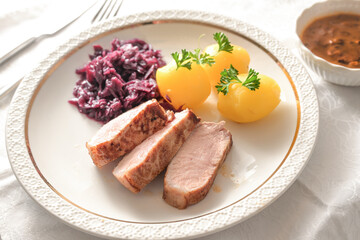Juicy roasted pork slices on a white plate with red cabbage, potatoes and gravy, hearty homemade meat dish for lunch or dinner, selected focus