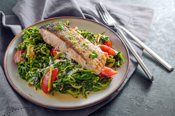 Juicy roasted salmon fillet with crispy skin on vegetables from cream spinach, onions and tomatoes, healthy fish meal with cutlery on a gray napkin and a slate surface