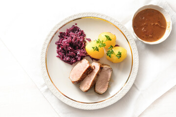 Slices of pork meat, red cabbage and potatoes with parsley garnish on a white plate and gravy in a bowl on a light table setting, high angle view from above, copy space