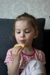 A small child or girl or baby sits on a sofa or armchair, holds a cookie in her hands, looks at it and chews. The child bites or eats the cookie. There are crumbs on her face.