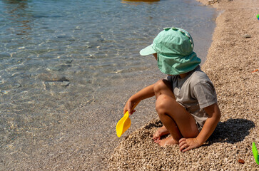 Little boy in a green cap playing with small pebbles on the seashore using a yellow spatula on a sunny day. Concept of childhood, outdoor play, and summer vacations by the sea.
