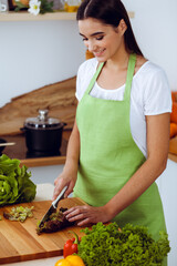 An attractive young dark-haired woman preparing delicious fresh vegetarian salad while standing and smiling near the kitchen desk. Cooking and householding concepts