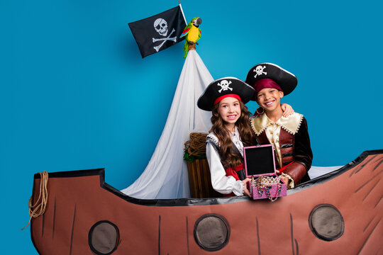 Portrait of two little kids halloween pirate costume embrace hold treasure sail boat isolated on blue color background