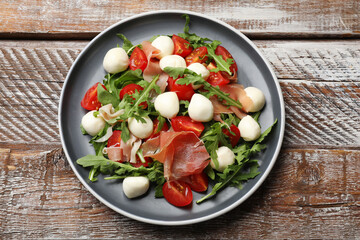 Tasty salad with arugula, mozzarella cheese, tomatoes and jamon on wooden table, top view