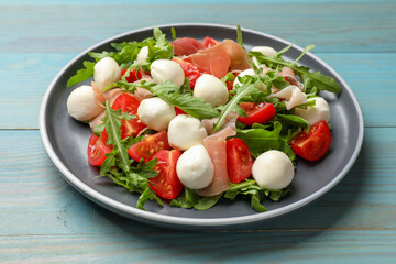 Tasty salad with arugula, mozzarella cheese, tomatoes and jamon on light blue wooden table, closeup