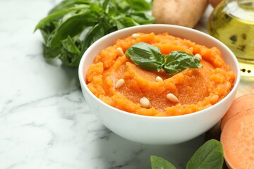 Tasty mashed sweet potato with basil, seeds in bowl and ingredients on white marble table, closeup