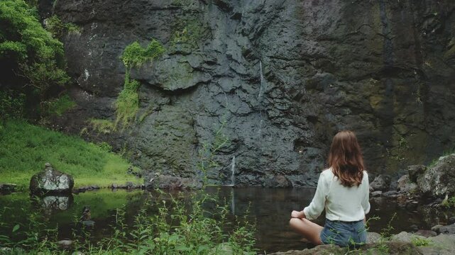 Young woman is practicing yoga and meditating near a small waterfall in a lush tropical forest. She is enjoying the peace and quiet of nature
