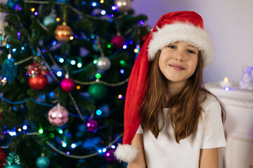 Close-up portrait of a smiling teenage girl wearing a Santa hat on the background of a Christmas tree