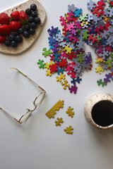 Colorful jigsaw puzzle pieces, cup of tea or coffee, eyeglasses and fresh berries for snacking. Hygge at home. Top view.