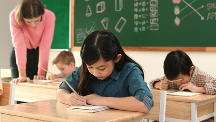 Attractive smart girl writing test or classwork while sitting at classroom. Diverse happy student taking notes and teacher help caucasian boy doing test while sitting in front of blackboard. Pedagogy.