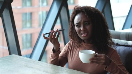 A very beautiful African American woman with long hair listens to a voice message on a smartphone and drinks tea or coffee while sitting at a table in a cafe in the dining room. The woman put her