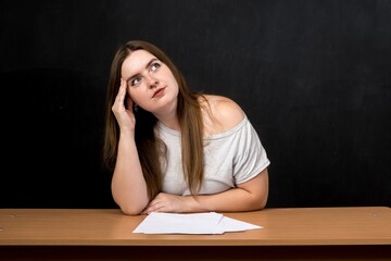 Young woman sitting at a desk, deep in thought with papers in front of her. She gazes upward,...