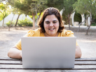 Woman with cognitive disability smiles and uses her laptop on the park. Lush greenery surrounds her.