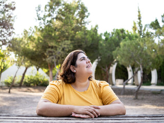 A woman with cognitive disability sits at a wooden table in a park, gazing upwards with a smile,...