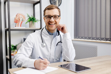 Portrait of young doctor wearing glasses and white uniform with stethoscope, speaking and consulting patient online, looking at camera, making video call, sitting at table in office