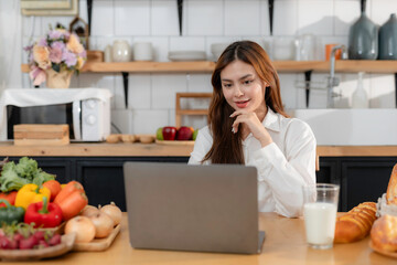 Young woman is using laptop, watching online cooking course and learning to prepare healthy food in her kitchen