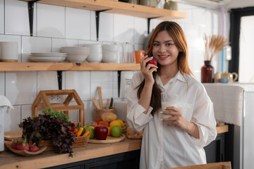 Young woman is holding a glass of milk and an apple, smiling in her kitchen with fresh fruits and vegetables on the counter