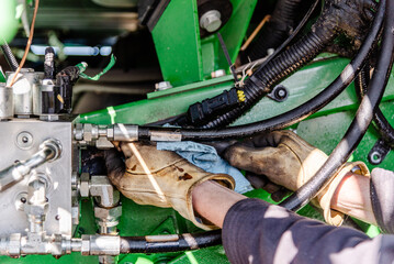 Farmer Mechanic Repairing Hydraulic Hose Under Combine Undercarriage, Wearing Yellow Gloves, Using Wrenches, Propped by Dual Tires, Agricultural Equipment Maintenance, Combine Harvester Repair