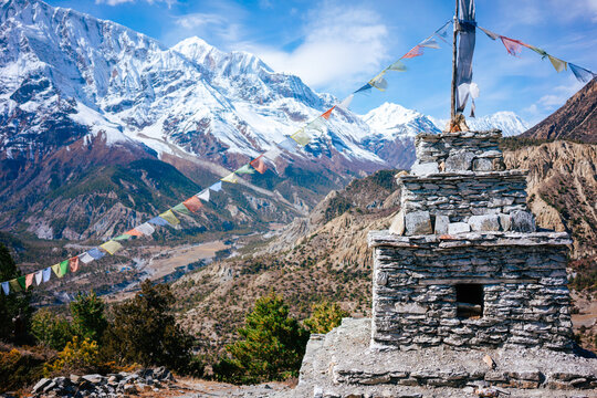 A stone chorten stands tall along the Annapurna Circuit, adorned with colorful prayer flags fluttering against a backdrop of dramatic snow-capped mountains in Nepal.